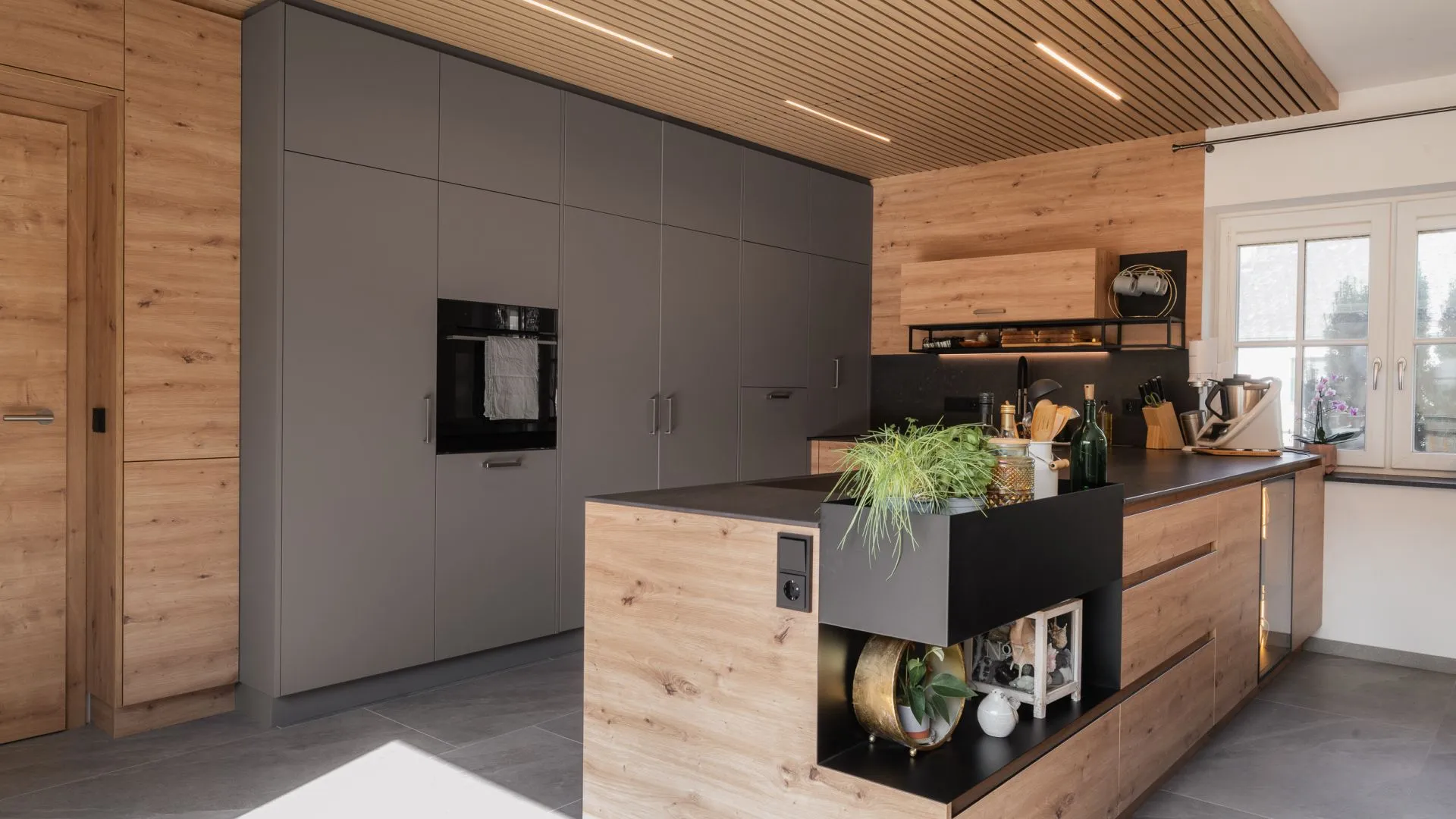 Moderne Küche mit warmen Holzfronten, dunklen Hochschränken und Arbeitsflächen sowie einer Akustikdecke mit integrierter Beleuchtung von HALBMAYR.