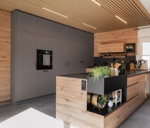Moderne Küche mit warmen Holzfronten, dunklen Hochschränken und Arbeitsflächen sowie einer Akustikdecke mit integrierter Beleuchtung von HALBMAYR.