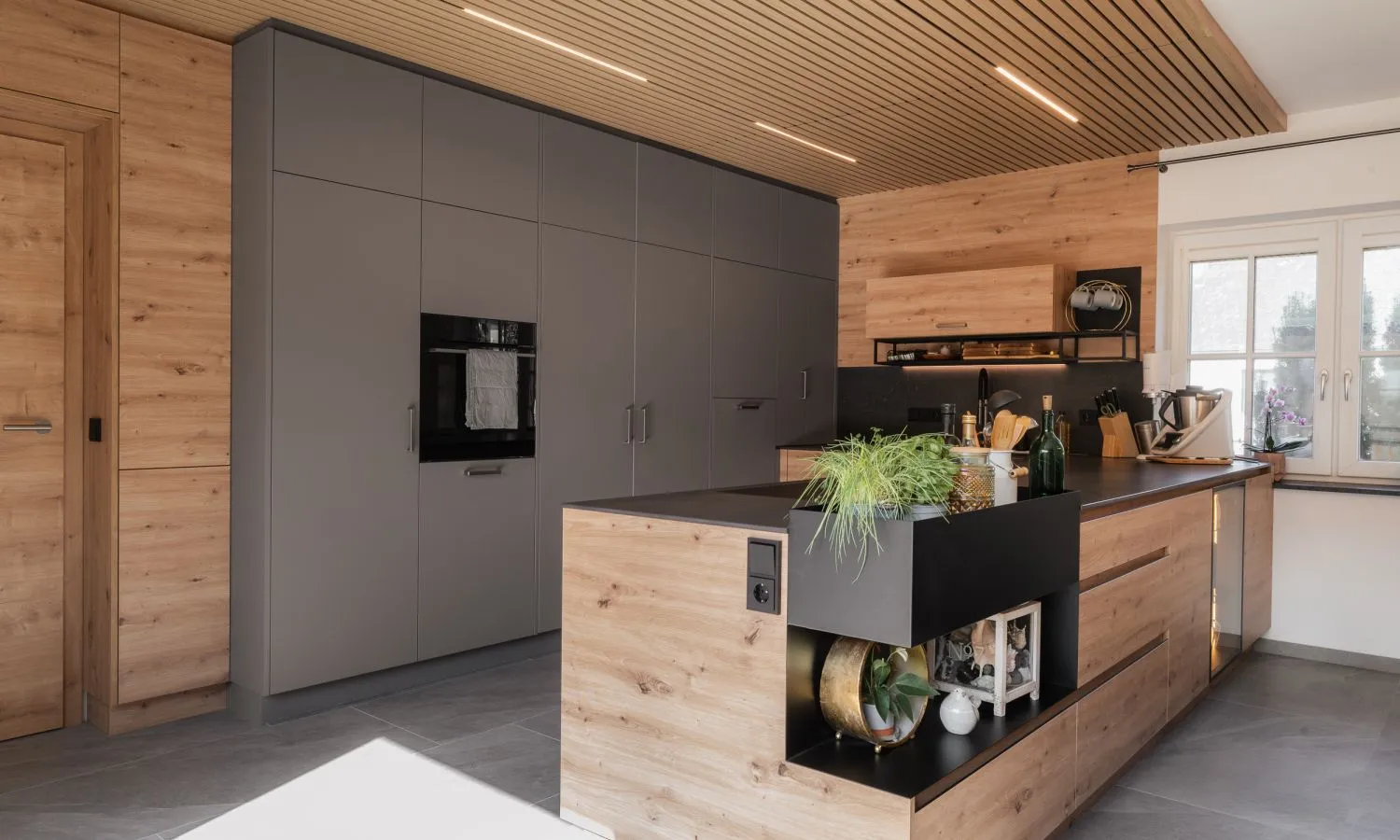 Moderne Küche mit warmen Holzfronten, dunklen Hochschränken und Arbeitsflächen sowie einer Akustikdecke mit integrierter Beleuchtung von HALBMAYR.
